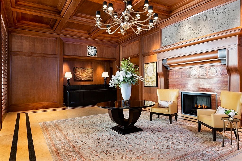 This interior shot showcases an elegant living room with rich wood paneling, a coffered ceiling, and a striking chandelier. A fireplace is flanked by comfortable armchairs, and a round table with a floral arrangement adds a touch of sophistication. The room exudes a sense of warmth and luxury, making it an inviting space for relaxation and conversation.