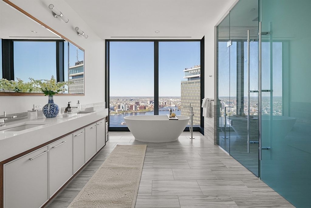 This is a luxurious primary bathroom featuring a modern freestanding bathtub positioned in front of a large window with a city view. The bathroom includes a long vanity with double sinks, sleek white cabinetry, and a glass-enclosed shower. The flooring is light gray tile, and a neutral-colored rug adds warmth to the space, creating a spa-like atmosphere.