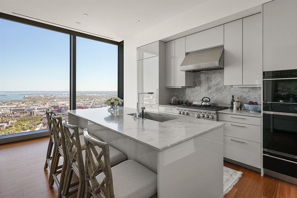 This is a modern kitchen with sleek, white cabinetry and stainless steel appliances. A large island with a marble countertop provides ample workspace and seating. The expansive window offers a stunning city view, enhancing the luxurious feel of the space. The camera is positioned to capture the entire kitchen, from the island to the window, creating a sense of depth and openness.