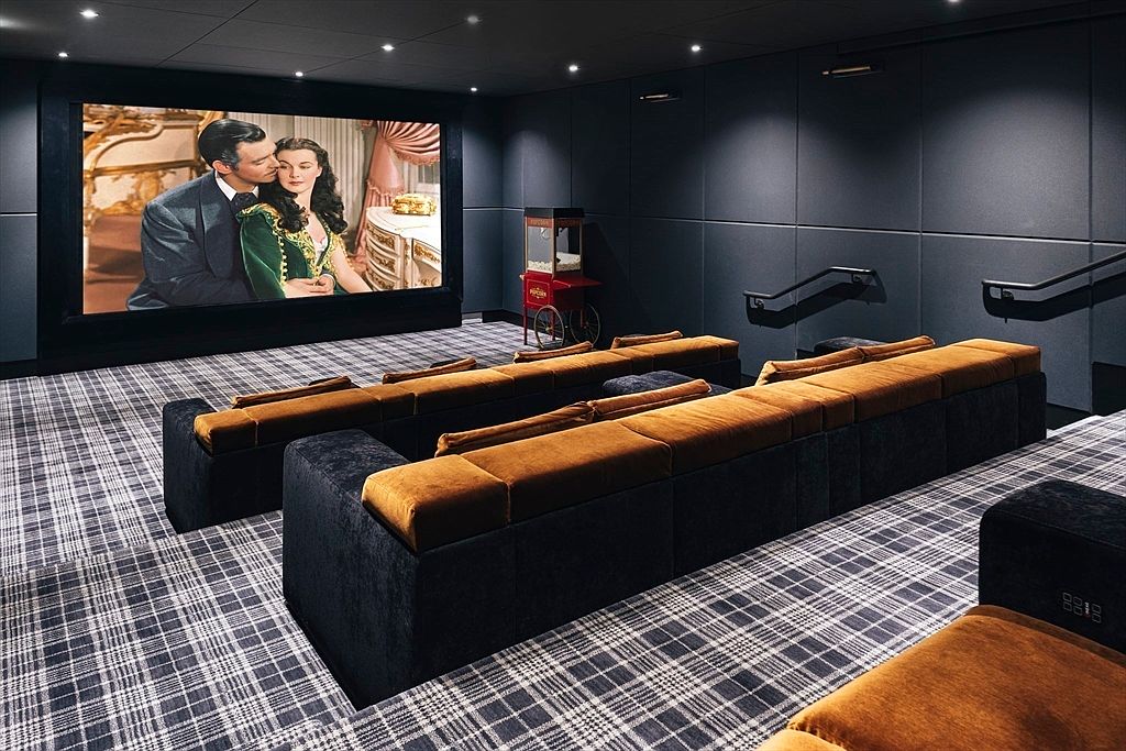 This is an interior shot of a home theater. The room features tiered seating with plush, dark-colored sofas and brown cushions, a large screen displaying a classic movie, and a popcorn machine in the background. The walls are dark, and the carpet has a plaid pattern, creating a cozy and immersive cinematic experience.