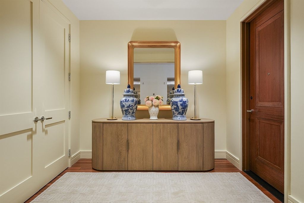This interior shot showcases a well-decorated hallway featuring a wooden cabinet with decorative vases and lamps, a large mirror, and a patterned rug. The walls are painted in a neutral tone, and there are two doors, one leading to another room and the other appearing to be a closet. The overall impression is elegant and inviting, suggesting a sophisticated living space.
