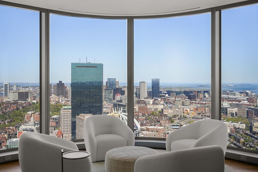 This interior shot showcases a modern living room with floor-to-ceiling curved windows offering a panoramic city view. Four white armchairs are arranged around a round ottoman, creating a cozy seating area. The room's design emphasizes the view, making it a focal point for potential buyers.