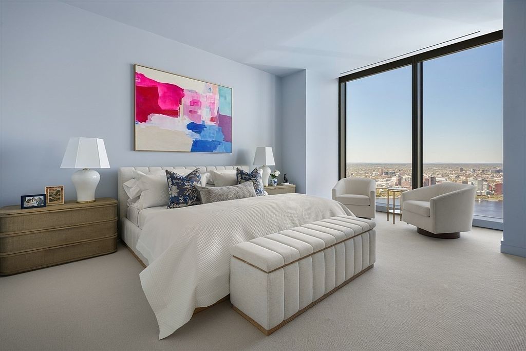 This is a bright and airy primary bedroom featuring a large bed with white linens and decorative pillows, complemented by a cushioned bench at the foot of the bed. An abstract painting adds a pop of color to the light blue walls, while floor-to-ceiling windows offer a stunning city view. The room is furnished with a wooden dresser and two armchairs, creating a serene and luxurious atmosphere.