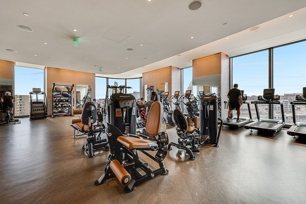 This is a well-equipped home gym featuring various weight machines, treadmills, and free weights. Large windows provide ample natural light and city views, enhancing the workout experience. The space is clean, modern, and designed for a comprehensive fitness routine.