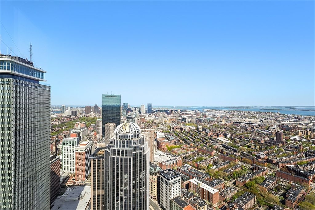This aerial view showcases a sprawling cityscape under a clear blue sky, dominated by modern skyscrapers and interspersed with residential areas featuring tree-lined streets. The architectural diversity is evident, with a mix of glass towers and traditional brick buildings, offering a sense of urban sophistication and vibrant community living. The perspective is a high-angle shot, emphasizing the scale and density of the city.