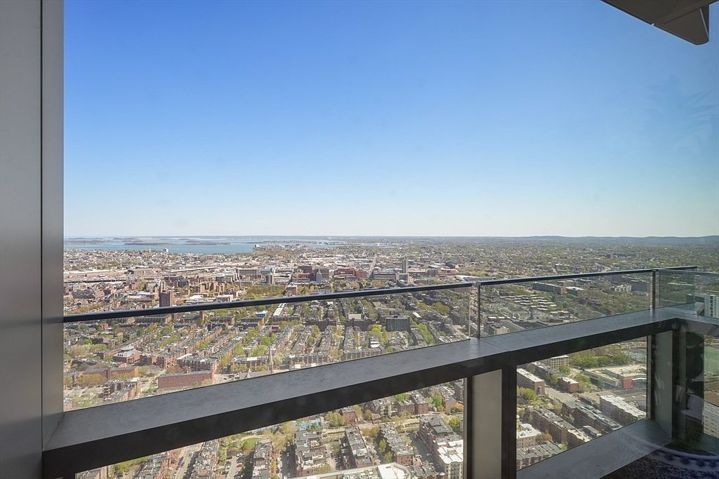 This aerial view showcases a sprawling cityscape under a clear blue sky, as seen from a high-rise balcony. The balcony features a modern glass railing and a concrete ledge, providing an unobstructed panorama of the urban landscape below. The perspective emphasizes the height and the expansive view, highlighting the property's prime location and stunning vistas.
