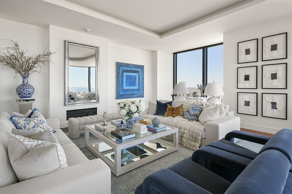 This is a beautifully decorated living room featuring a neutral color palette with pops of blue. The room includes two sofas, a mirrored coffee table, and a large window offering natural light. Artwork and decorative items add a touch of elegance and sophistication to the space.