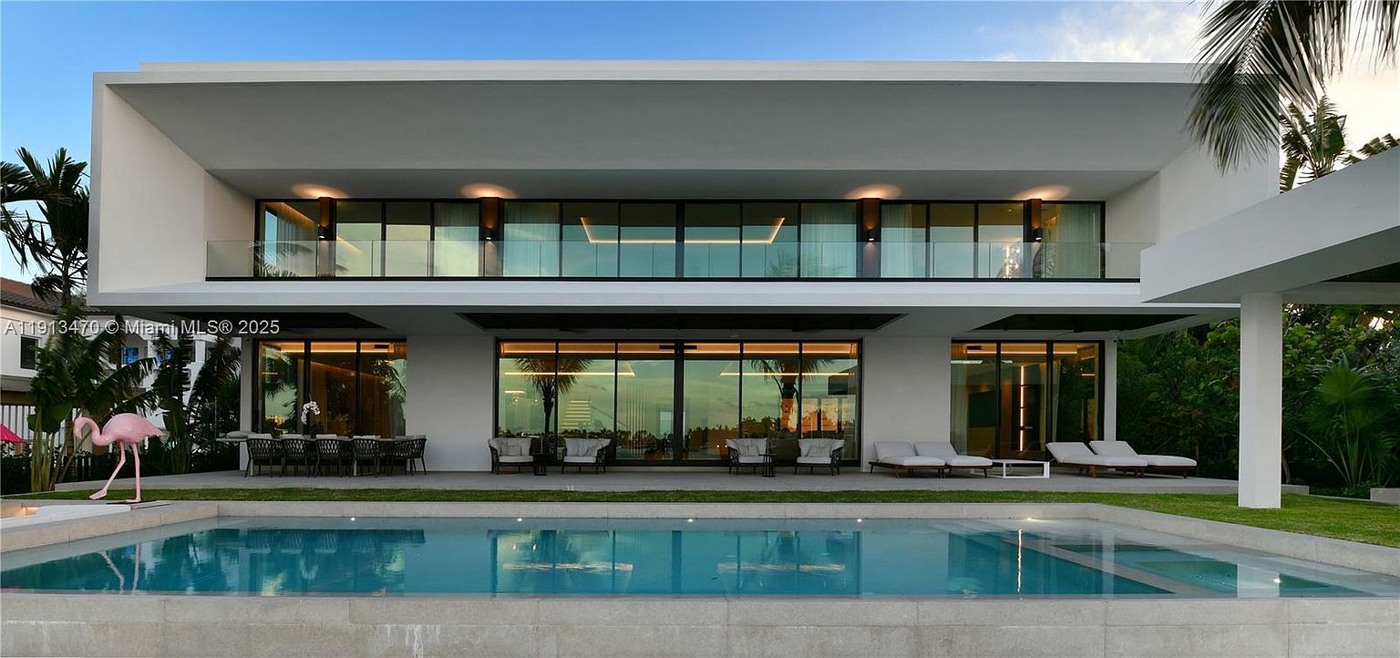This modern, two-story luxury residence features a clean, minimalist aesthetic with crisp white exterior walls and extensive glass glazing. The rear of the home opens onto a spacious covered patio area, which transitions seamlessly into a rectangular swimming pool. The property is surrounded by lush tropical landscaping, creating a private and serene outdoor living environment.