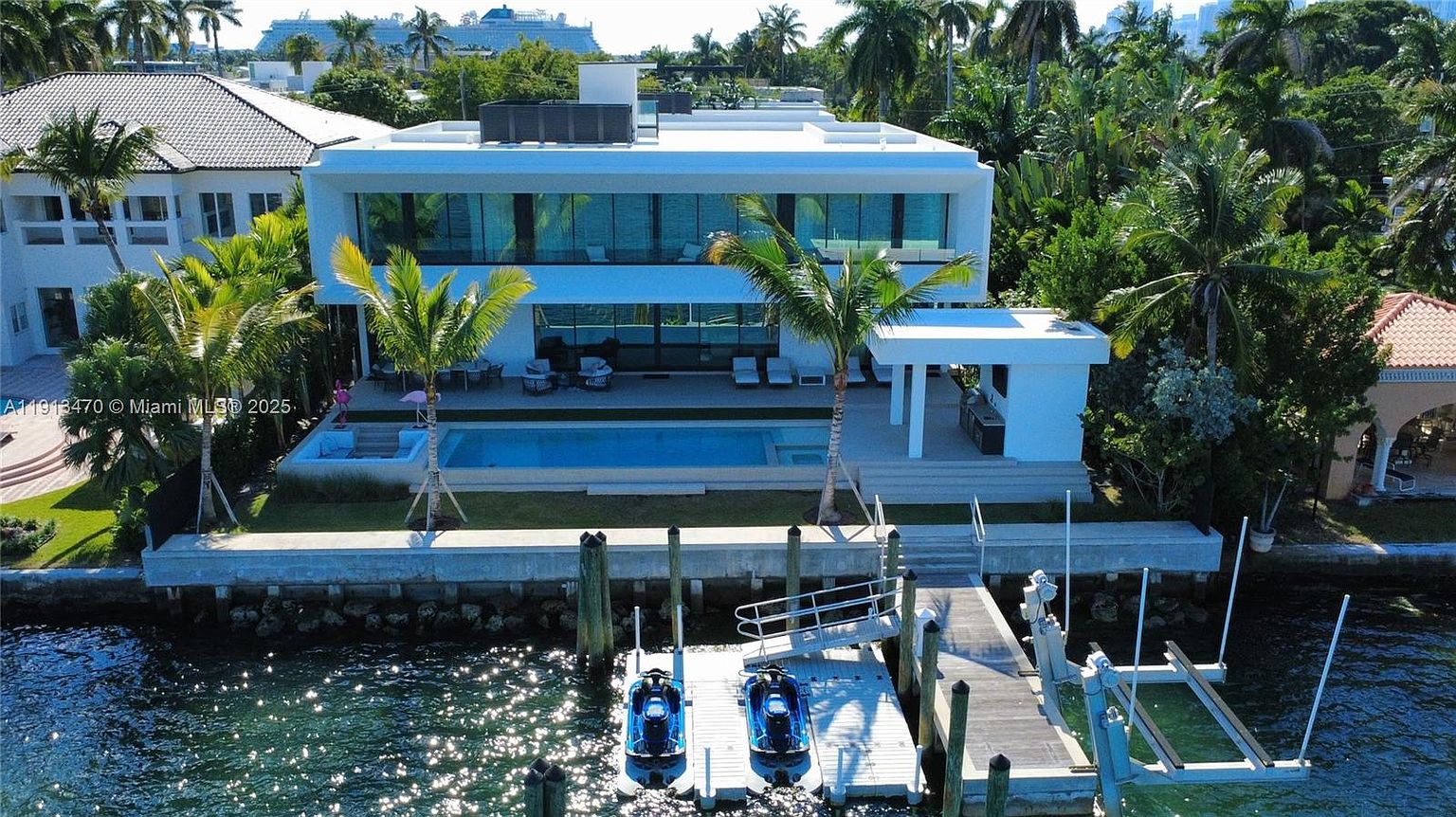This aerial perspective captures a modern, two-story waterfront residence featuring a sleek white facade and expansive glass walls. The property includes a private swimming pool, a covered outdoor lounge area, and a private dock equipped with two jet skis extending into the waterway. Lush tropical landscaping and palm trees frame the home, emphasizing its luxurious coastal setting.