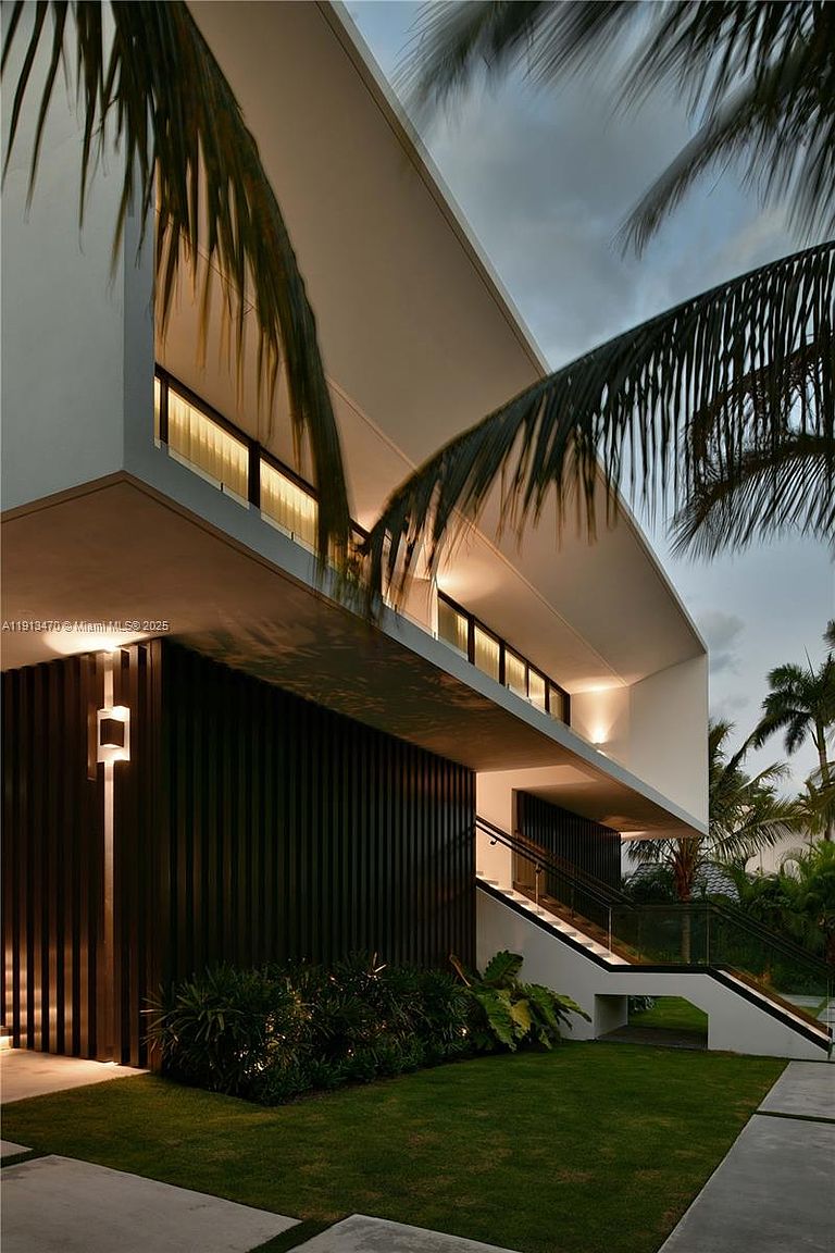 This striking modern residence features a dramatic, curved roofline and a sophisticated exterior palette of dark vertical slats and crisp white surfaces. A set of illuminated stairs with a glass railing leads to the elevated main level, while lush tropical landscaping and palm trees frame the structure. The scene is captured at dusk, with warm accent lighting highlighting the architectural textures and creating an inviting, high-end atmosphere.