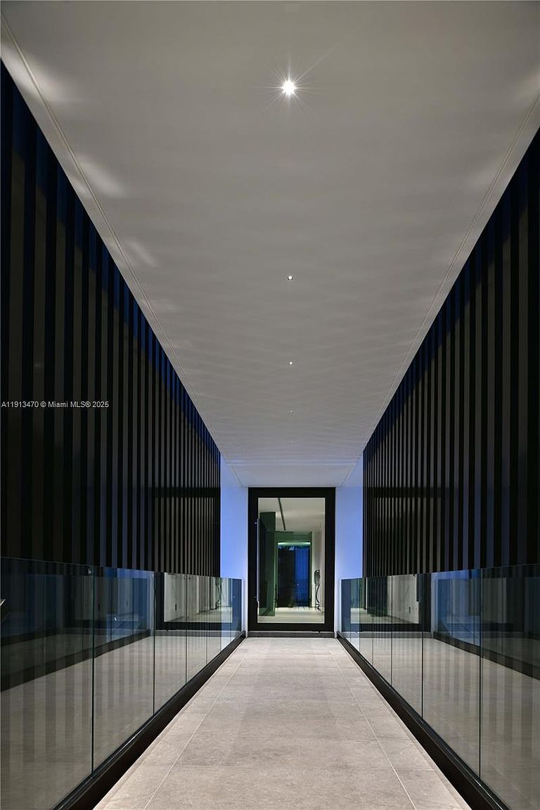 This sleek, modern hallway features a long, symmetrical corridor defined by dark, vertical-slatted walls and glass railings on either side. The perspective draws the eye toward a central doorway at the end, which is illuminated by a soft blue glow. The neutral-toned tile flooring and recessed ceiling lighting create a minimalist and sophisticated atmosphere.