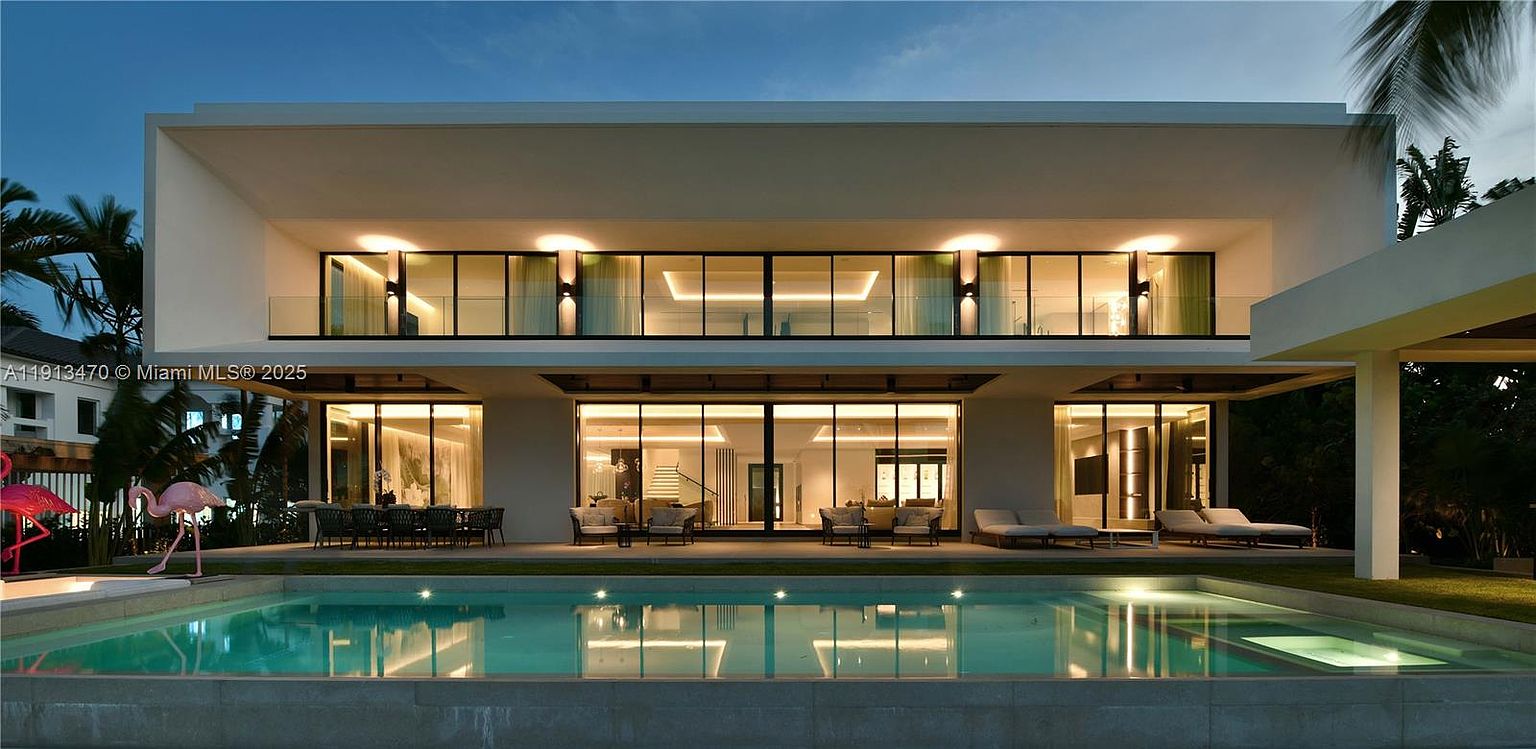 This modern, two-story luxury residence features a symmetrical design with extensive glass walls that provide a seamless transition between the interior living spaces and the outdoor patio. A large, rectangular swimming pool sits in the foreground, reflecting the clean lines of the architecture and the warm glow of the recessed lighting. The property is framed by lush tropical landscaping, including palm trees and decorative flamingo statues, creating a sophisticated and serene resort-like atmosphere.