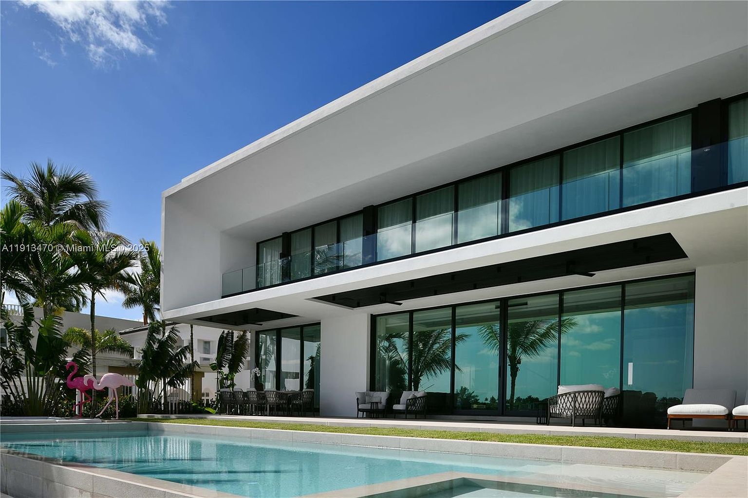 This modern, two-story luxury residence features a sleek white facade with expansive floor-to-ceiling glass walls that seamlessly connect the interior living spaces to the outdoor patio. A pristine, rectangular swimming pool sits in the foreground, reflecting the bright blue sky and the surrounding palm trees. The patio area is furnished with contemporary outdoor seating and dining sets, creating an inviting space for relaxation and entertaining.