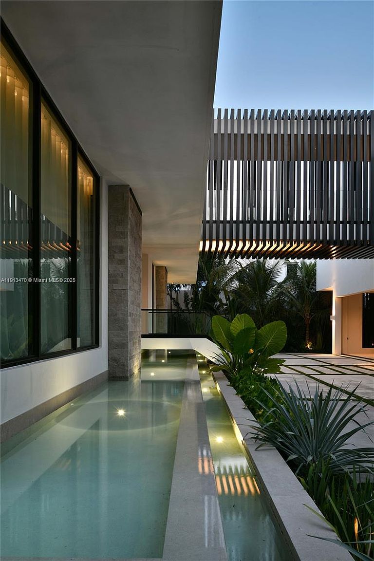 This modern architectural exterior features a sleek, narrow reflecting pool that runs alongside a glass-walled structure. A striking overhead slatted wood canopy creates dramatic linear shadows, while lush tropical landscaping and integrated lighting enhance the sophisticated, resort-like atmosphere. The perspective captures the clean lines and high-end design of the property's outdoor living space.