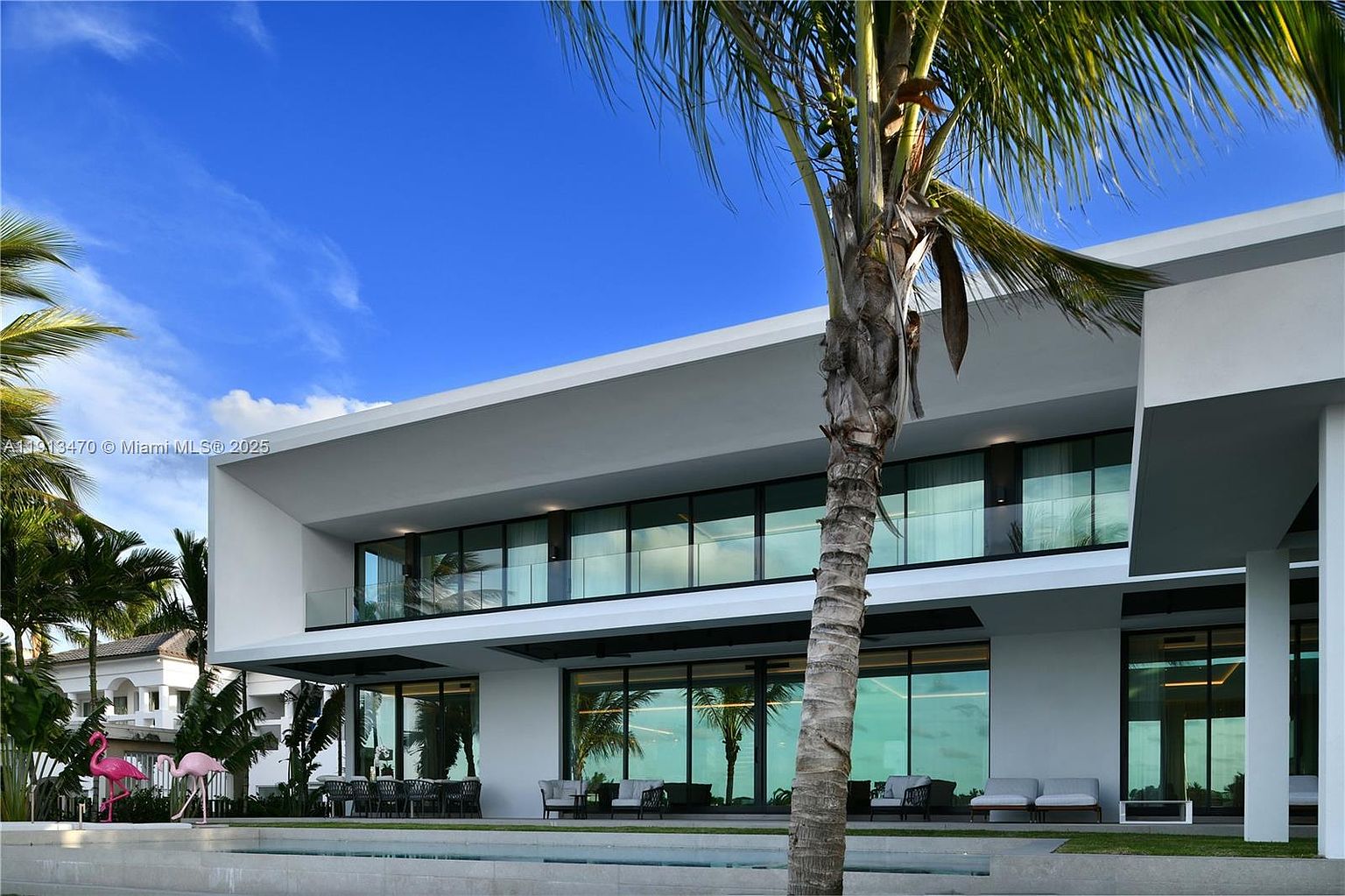 This modern, two-story luxury residence features a sleek, minimalist design with expansive glass walls and a deep, cantilevered roofline. The rear exterior showcases a spacious patio area furnished with outdoor seating, overlooking a pristine swimming pool. A prominent palm tree stands in the foreground, framing the architectural structure against a clear blue sky.