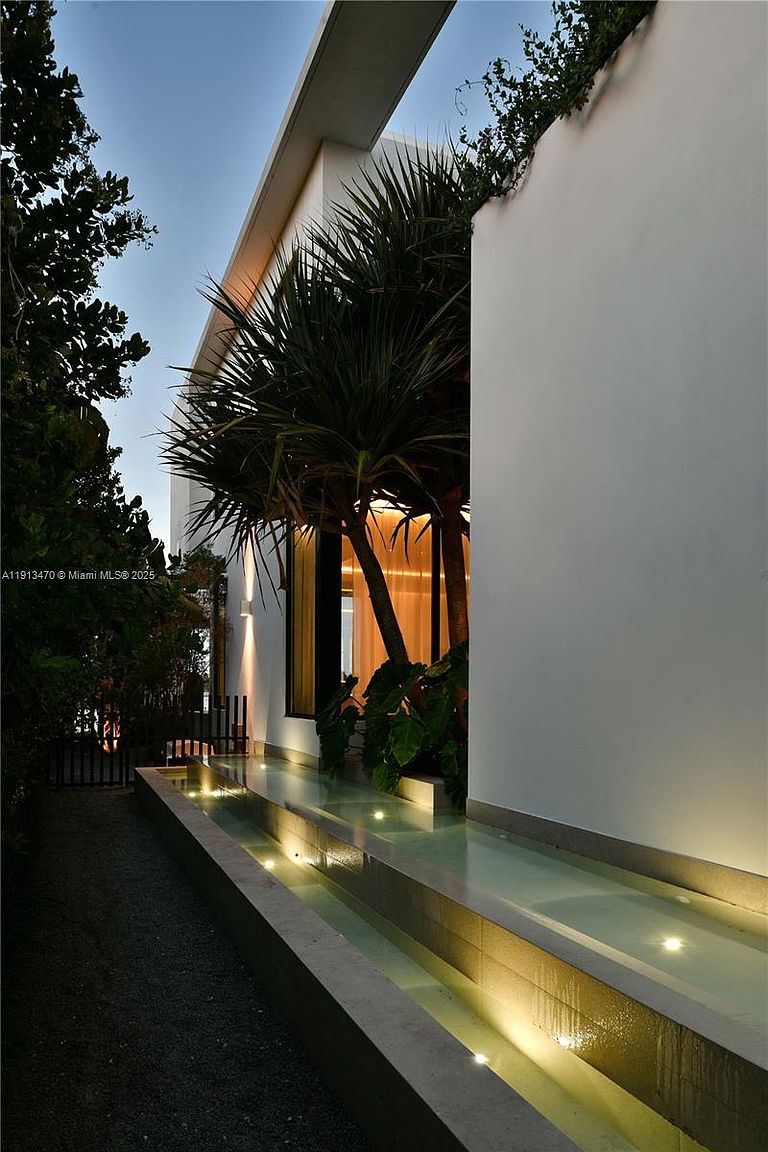 This side view captures a modern architectural design featuring a long, illuminated water feature running parallel to a smooth, white exterior wall. A lush, tropical palm tree stands prominently near the structure, casting dramatic shadows against the wall, while warm interior lighting glows through tall windows. The perspective is low and cinematic, emphasizing the sleek lines and serene, resort-like atmosphere of the property's exterior.