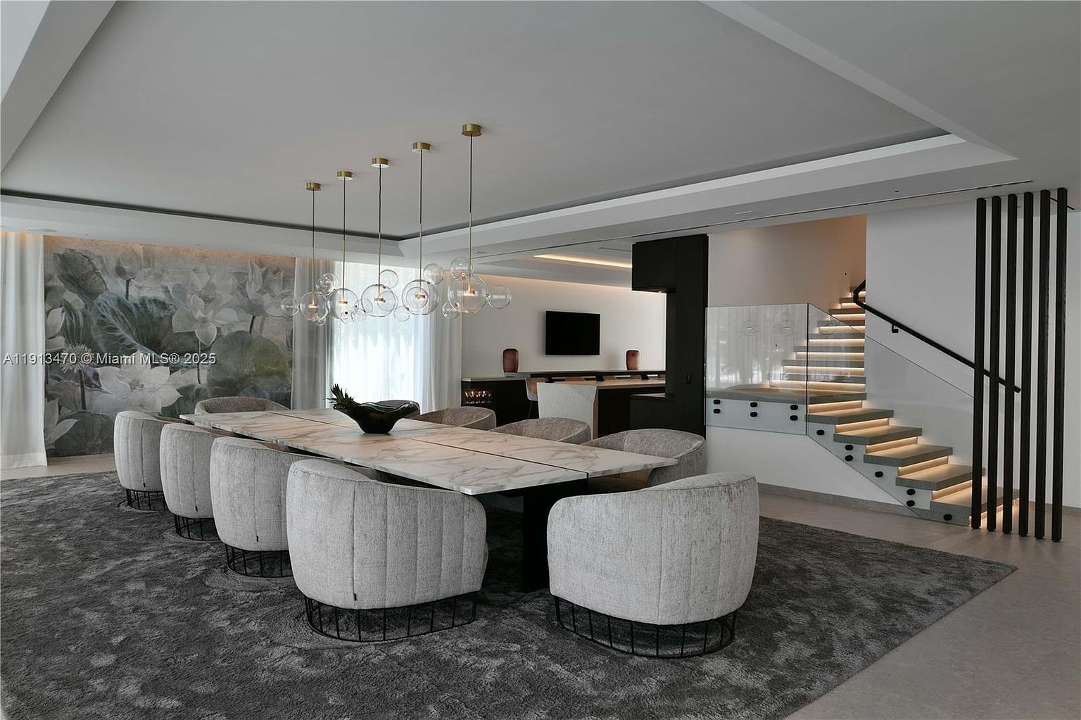 This sophisticated dining area features a long, light-toned stone table surrounded by eight plush, curved-back chairs set atop a textured gray area rug. A striking cluster of spherical glass pendant lights hangs above the table, while a large, artistic mural depicting lotus flowers serves as a dramatic backdrop. To the right, a modern staircase with glass railings and integrated step lighting leads to an upper level, contributing to the home's contemporary and luxurious aesthetic.
