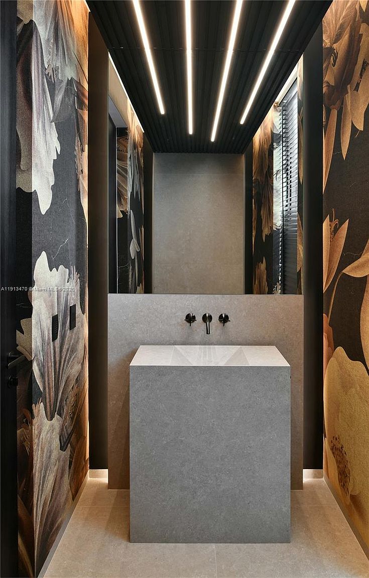 This sophisticated powder room features a striking, minimalist stone pedestal sink centered against a neutral stone wall. The space is framed by dramatic, dark floral wallpaper on both side walls, creating a moody and luxurious atmosphere. Overhead, a slatted ceiling with integrated linear lighting adds a modern architectural touch, while a large mirror reflects the depth of the room.