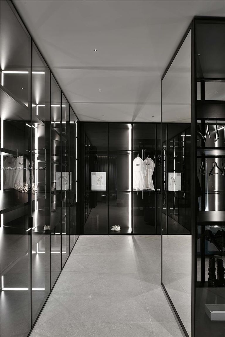 This sophisticated walk-in closet features a sleek, modern design with floor-to-ceiling glass-front cabinetry and integrated vertical LED lighting. The space is finished with large-format neutral floor tiles and a monochromatic color palette, creating a high-end, boutique-like atmosphere. The perspective is centered, looking down the main aisle between the mirrored and glass-enclosed storage units.