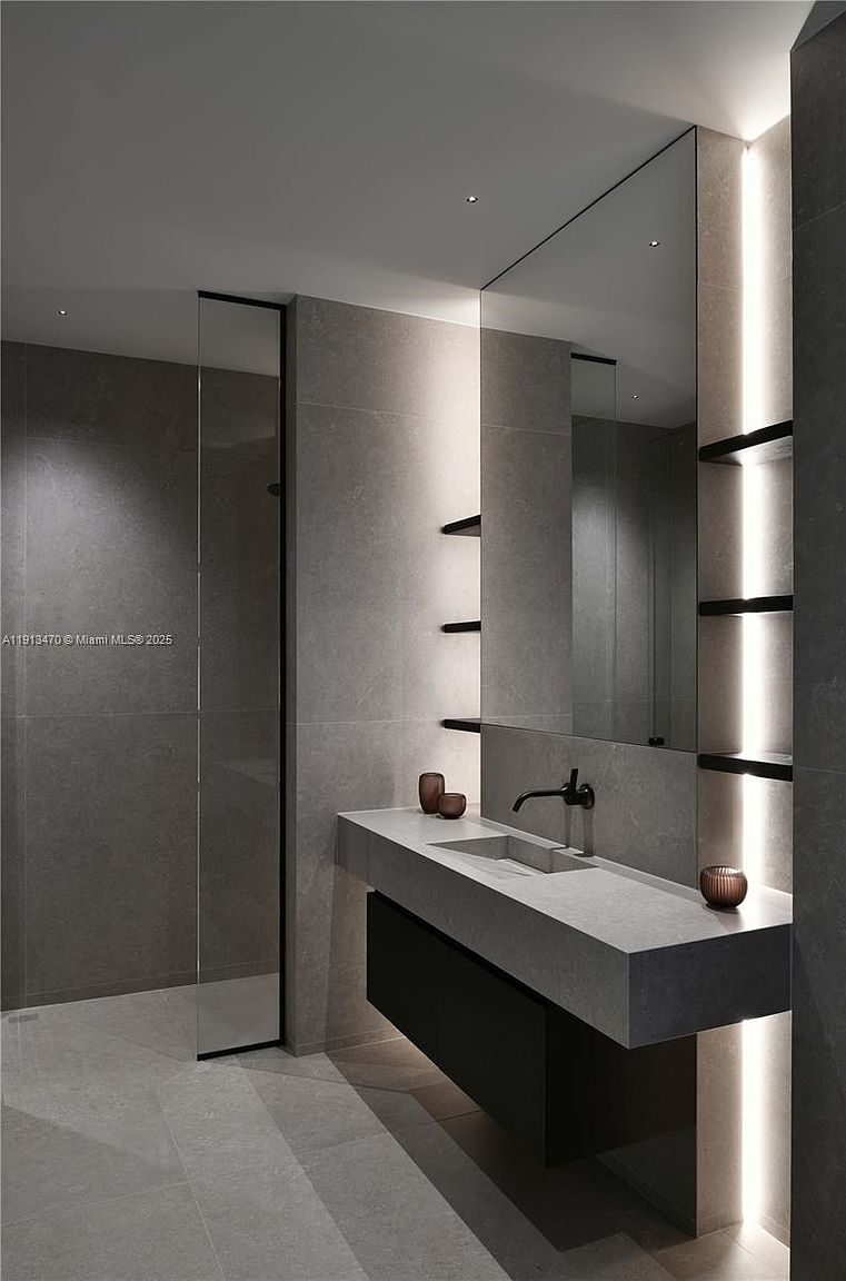 This modern, minimalist bathroom features a floating stone vanity with a sleek, dark-toned base and a matte black faucet. The space is defined by large-format grey stone tiles, a frameless glass shower enclosure, and integrated vertical LED lighting that highlights the shelving and mirror area. The overall aesthetic is sophisticated and contemporary, emphasizing clean lines and a monochromatic color palette.