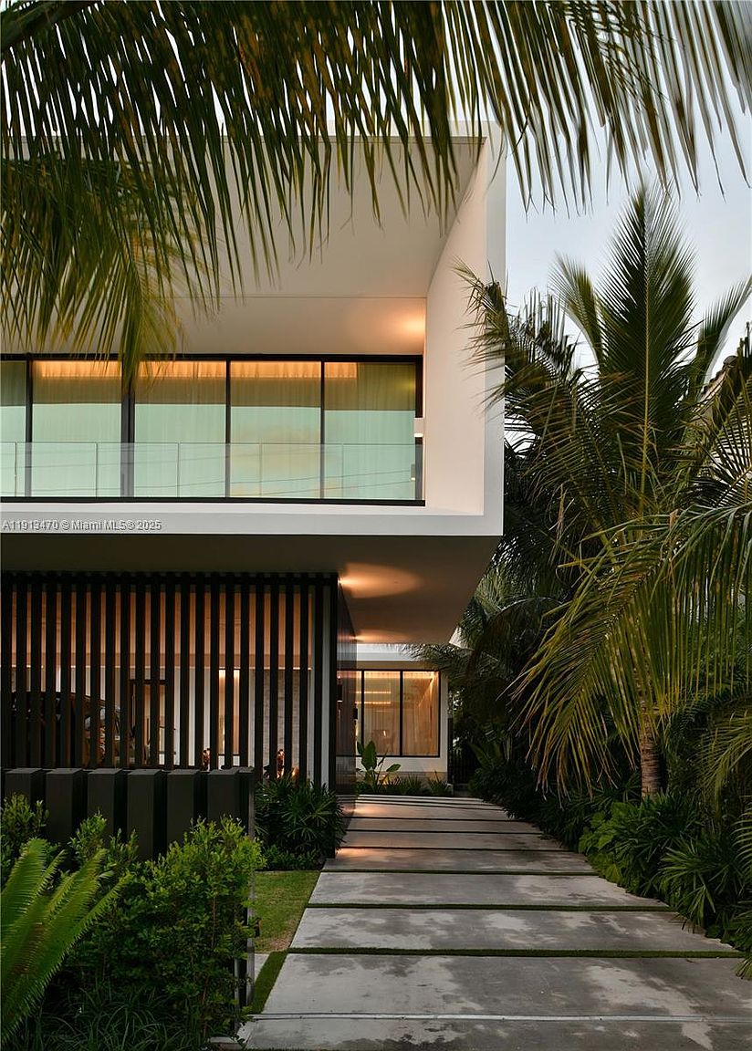 This modern architectural residence features a striking entryway defined by a concrete paver walkway leading toward a dramatic, vertical slat screen. The upper level showcases a clean, minimalist white facade with large windows that span the width of the structure. Lush tropical landscaping, including mature palm trees, frames the home, creating a sophisticated and serene atmosphere.