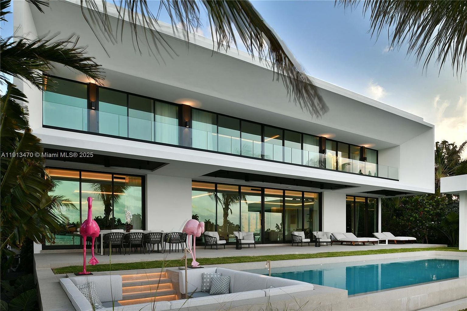 This modern luxury residence features a striking two-story architectural design with expansive glass walls and a sleek, minimalist aesthetic. The rear exterior showcases a spacious patio area, a sunken lounge space adjacent to a pristine swimming pool, and whimsical flamingo sculptures that add a playful touch to the manicured lawn. The composition captures the seamless transition between indoor and outdoor living spaces, emphasizing the home's resort-like atmosphere.
