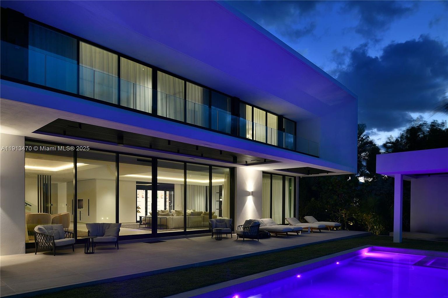 This modern luxury residence is captured at twilight, showcasing a sleek architectural design with expansive glass walls that provide a seamless transition between the interior living space and the outdoor patio. The scene features a pristine swimming pool with vibrant purple underwater lighting, reflecting the contemporary aesthetic of the home. A spacious covered patio area is furnished with comfortable lounge seating, creating an inviting atmosphere for evening relaxation and entertainment.