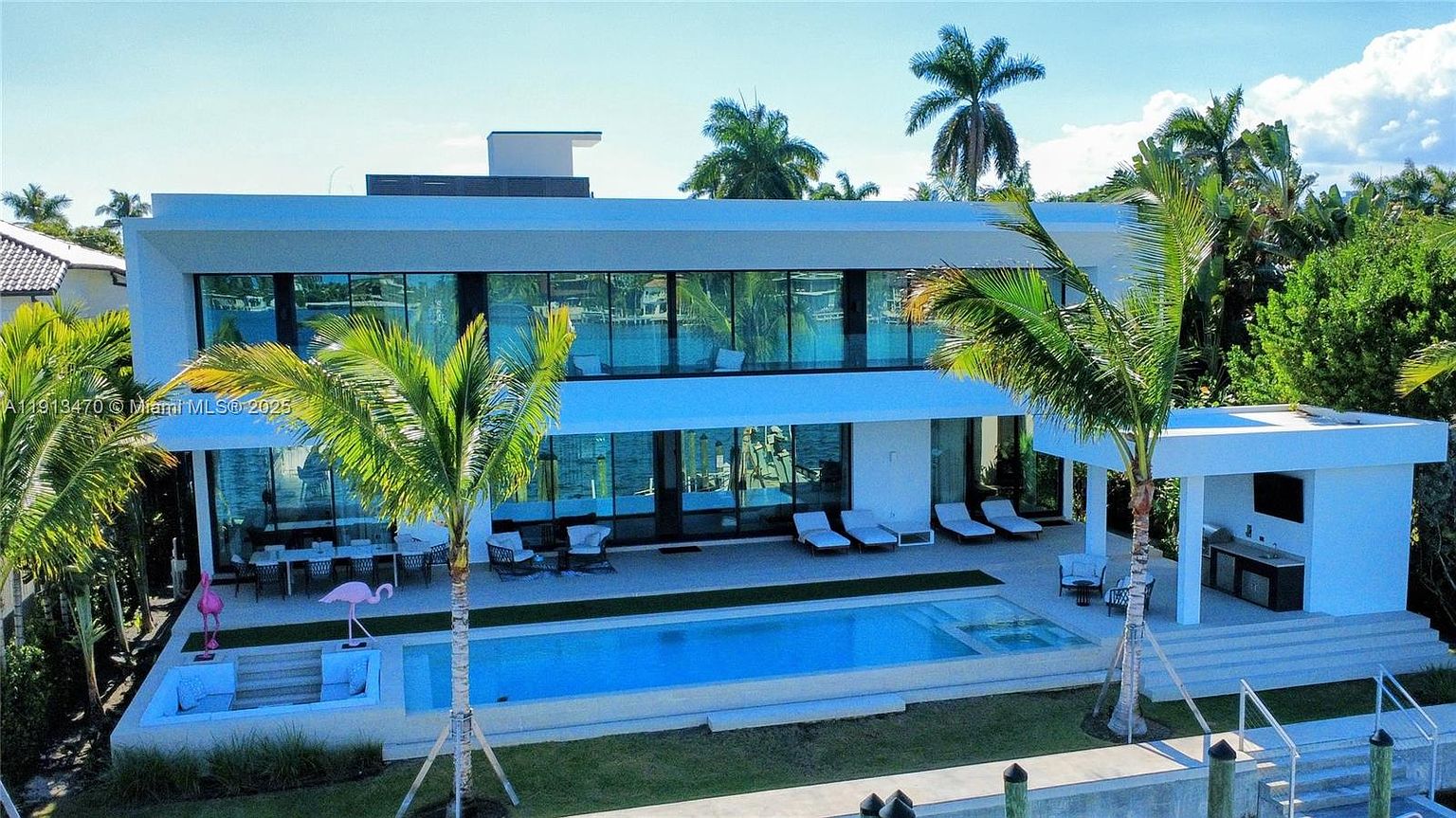 This modern, two-story waterfront residence features a sleek, minimalist design with extensive glass walls that offer panoramic views of the water. The rear exterior includes a spacious patio area, a rectangular swimming pool with an integrated spa, and a covered outdoor kitchen space. Lush palm trees and manicured landscaping frame the property, creating a luxurious and serene tropical atmosphere.