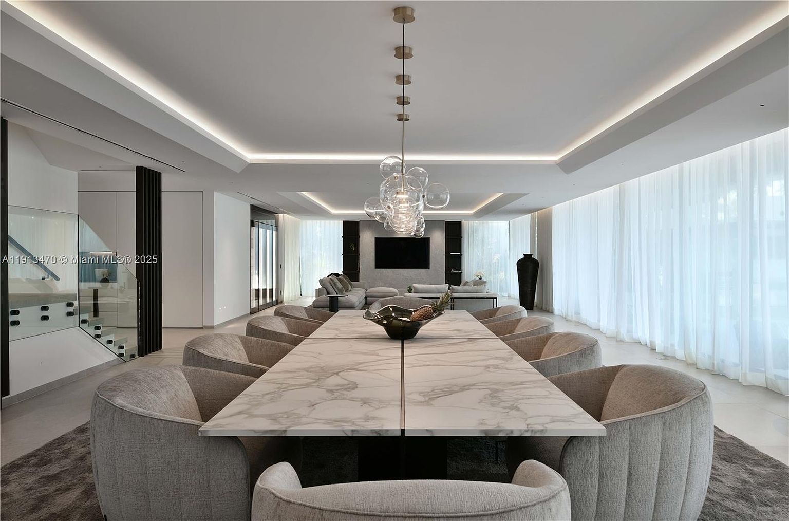 This sophisticated dining area features a long, light-toned stone table surrounded by ten upholstered chairs, centered beneath a modern glass bubble chandelier. The room is designed with a minimalist aesthetic, incorporating recessed ceiling lighting, soft white drapery covering large windows, and a neutral color palette. In the background, a spacious living area with plush seating and a wall-mounted television completes the open-concept layout, creating an atmosphere of refined luxury.