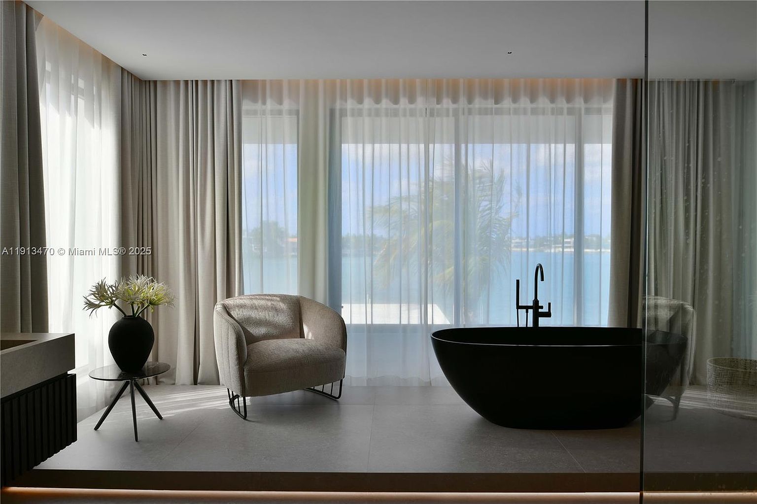 This luxurious primary bathroom features a sleek, matte black freestanding soaking tub positioned as a focal point against a backdrop of floor-to-ceiling windows. A contemporary, curved armchair sits beside a small glass-topped side table, creating a serene lounge area within the space. The room is bathed in soft, natural light filtered through sheer curtains, offering a tranquil view of the water beyond.