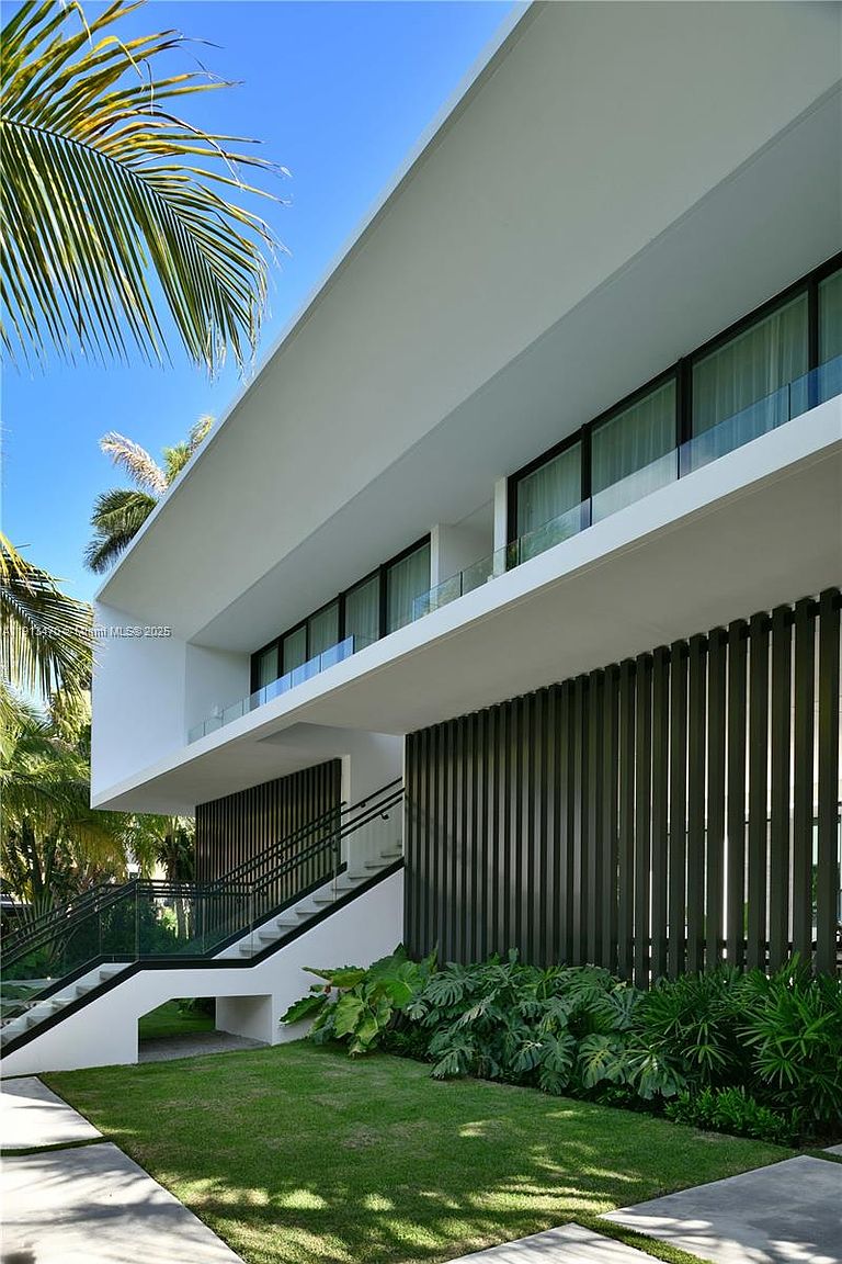 This modern architectural exterior features a striking cantilevered upper level with floor-to-ceiling glass walls, creating a sleek and minimalist aesthetic. A staircase with glass railings leads up to the elevated living space, while the ground level is accented by vertical dark slats and lush tropical landscaping. The bright, sunny perspective highlights the clean lines and sophisticated design of this contemporary residence.