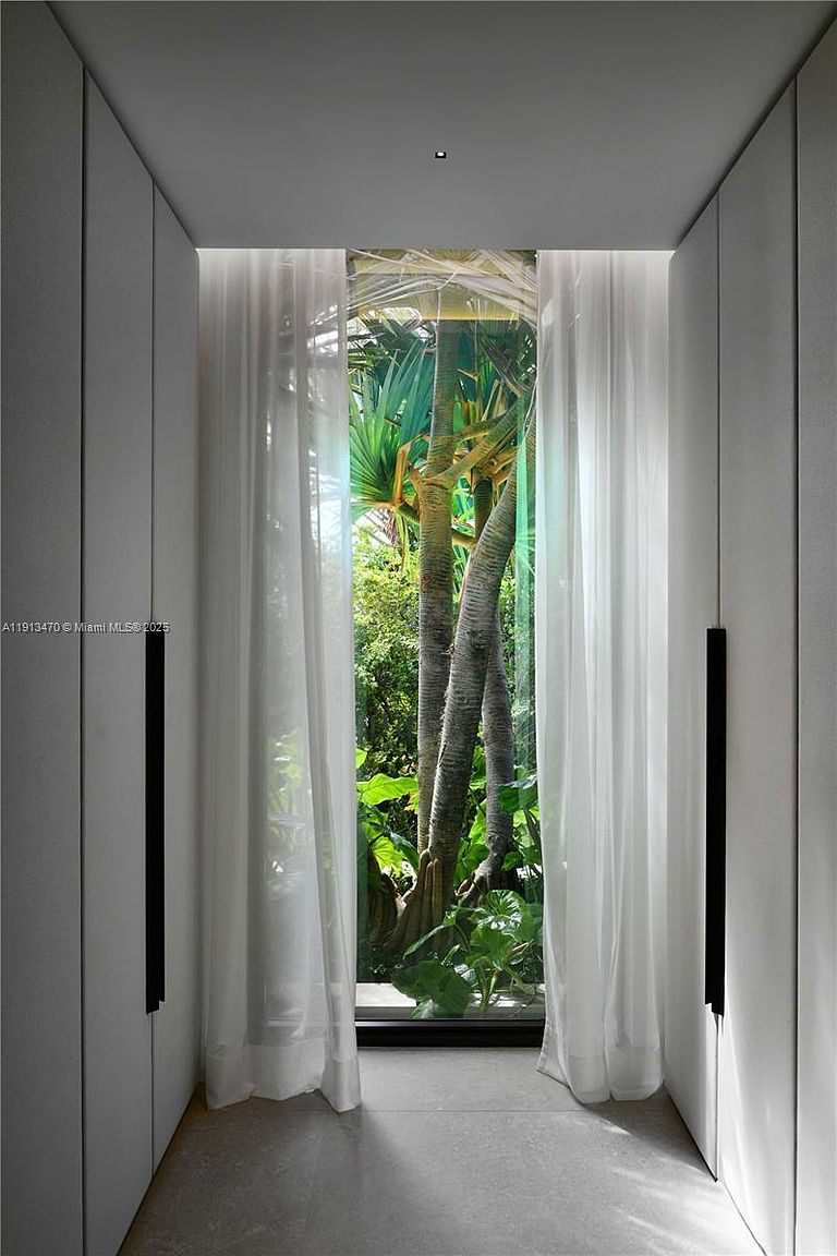 This sophisticated walk-in closet features minimalist, full-height cabinetry with sleek black vertical hardware on both sides of the corridor. At the end of the hallway, a large floor-to-ceiling window provides a serene, direct view of lush tropical greenery and tree trunks, blurring the line between indoor and outdoor spaces. The space is framed by soft, sheer white curtains that add a touch of elegance and texture to the clean, modern aesthetic.