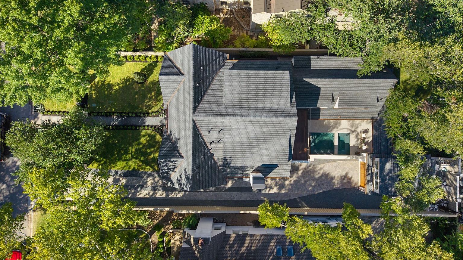 This aerial shot showcases a well-maintained property featuring a gray shingle roof, lush green lawns, and mature trees providing ample shade. A rectangular pool is visible in the backyard, complemented by a stone patio and outdoor seating. The overall impression is one of a private and luxurious residential estate.