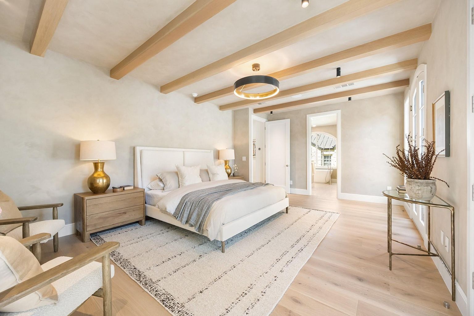 This is a bright and airy primary bedroom featuring a neutral color palette with wooden ceiling beams. The room is furnished with a white upholstered bed, a light wood nightstand with a gold lamp, and a glass-topped side table with a potted plant. A large area rug anchors the space, creating a cozy and inviting atmosphere.