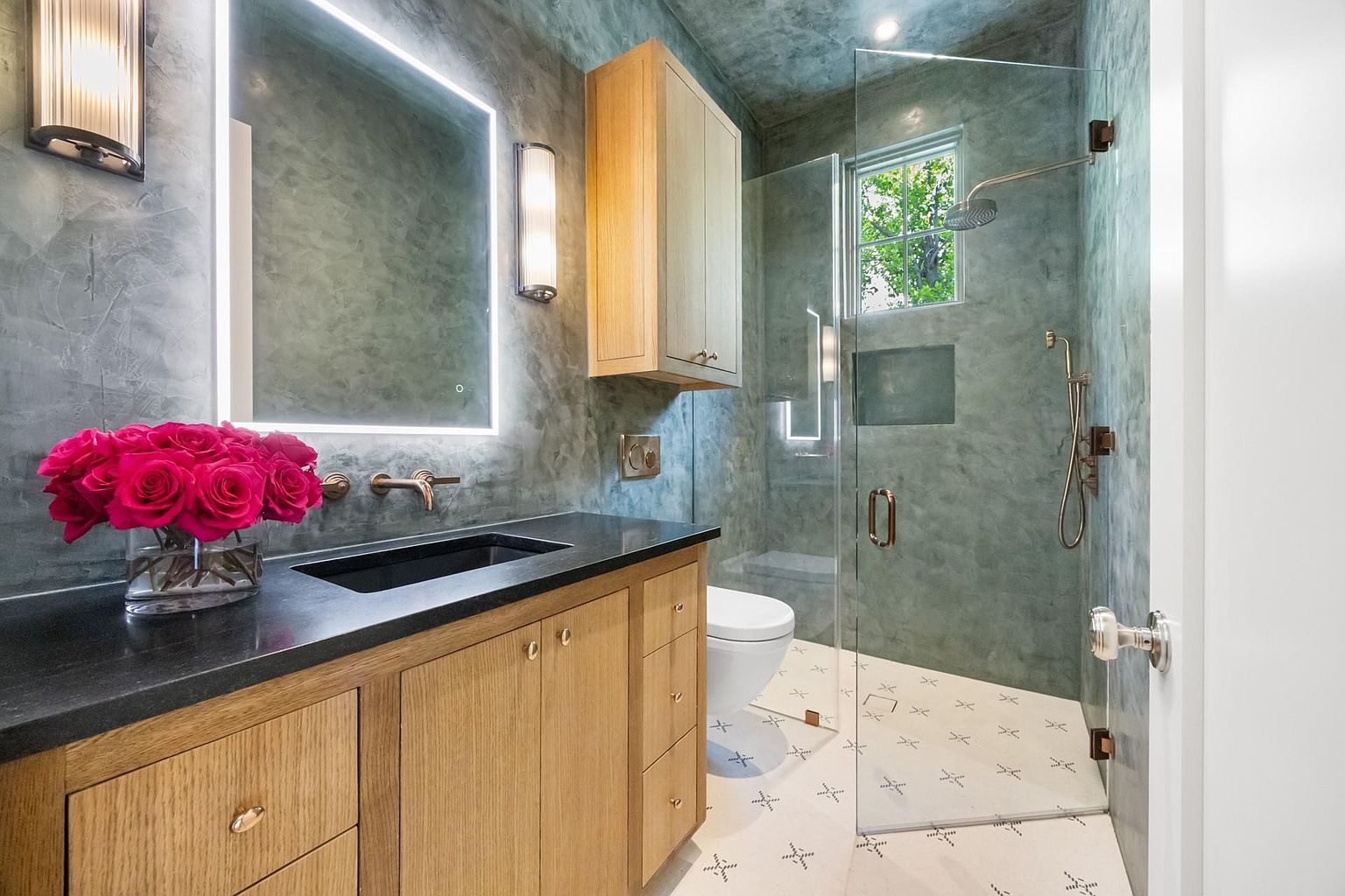 This is a modern bathroom featuring a vanity with a dark countertop and light wood cabinetry, complemented by a square, illuminated mirror and stylish sconces. The shower is enclosed with glass and showcases a rainfall showerhead, while the floor is adorned with patterned tiles. The overall design exudes a contemporary and luxurious feel.