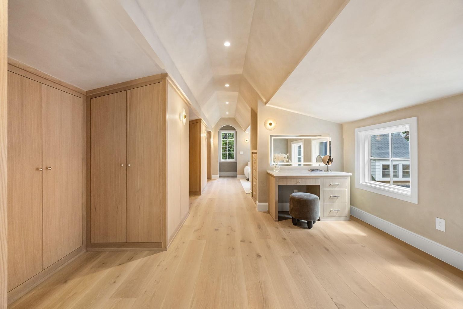 This is a well-lit walk-in closet featuring custom wooden cabinetry and hardwood flooring. The closet includes a vanity area with a mirror and stool, as well as a window providing natural light. The overall impression is one of luxury and organization.