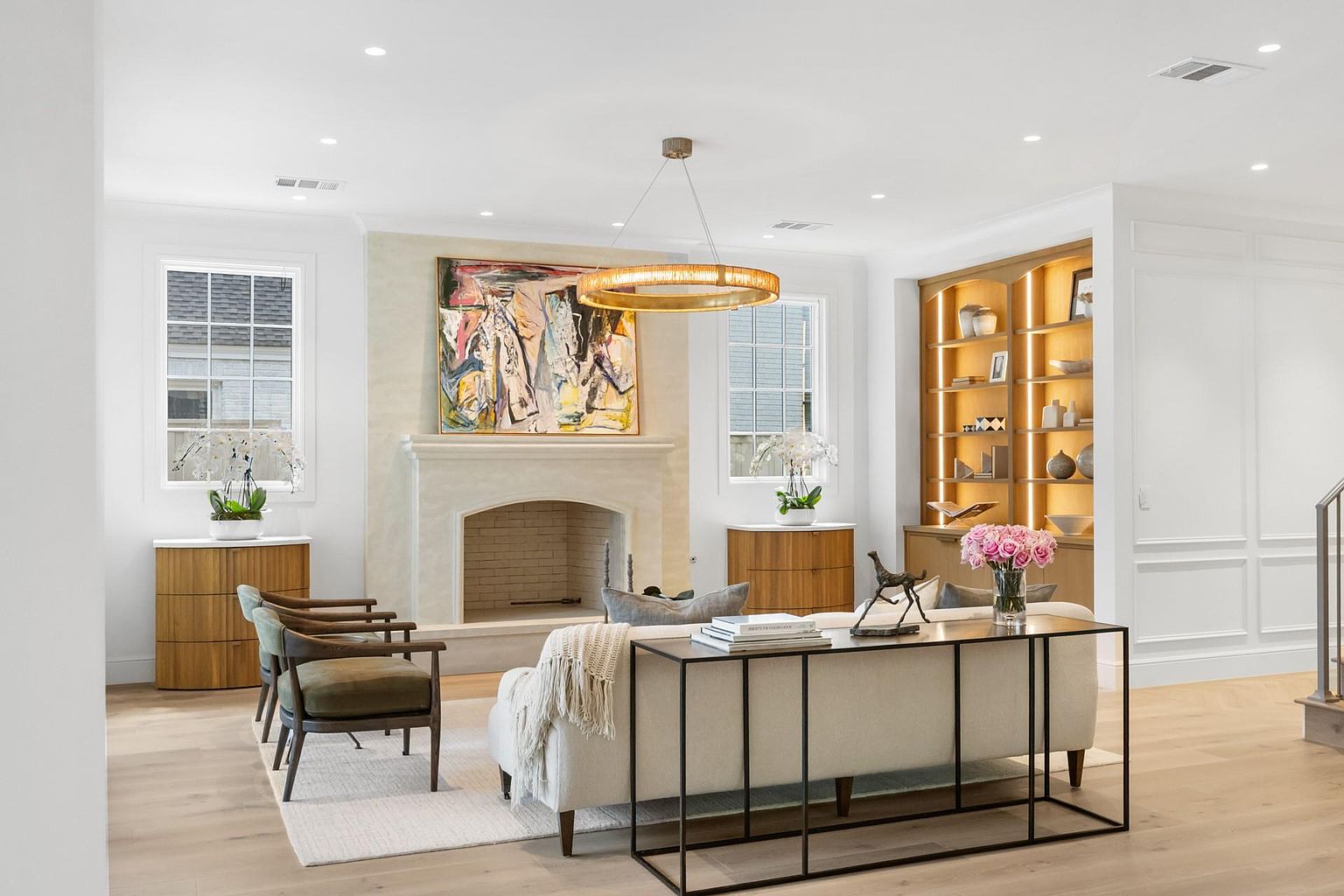 This is an interior shot of a well-lit living room featuring a fireplace with an abstract painting above it, two armchairs, and a sofa with a console table behind it. The room has hardwood floors, built-in shelving with decorative items, and large windows that provide natural light. The overall style is elegant and modern, creating a welcoming and sophisticated atmosphere.