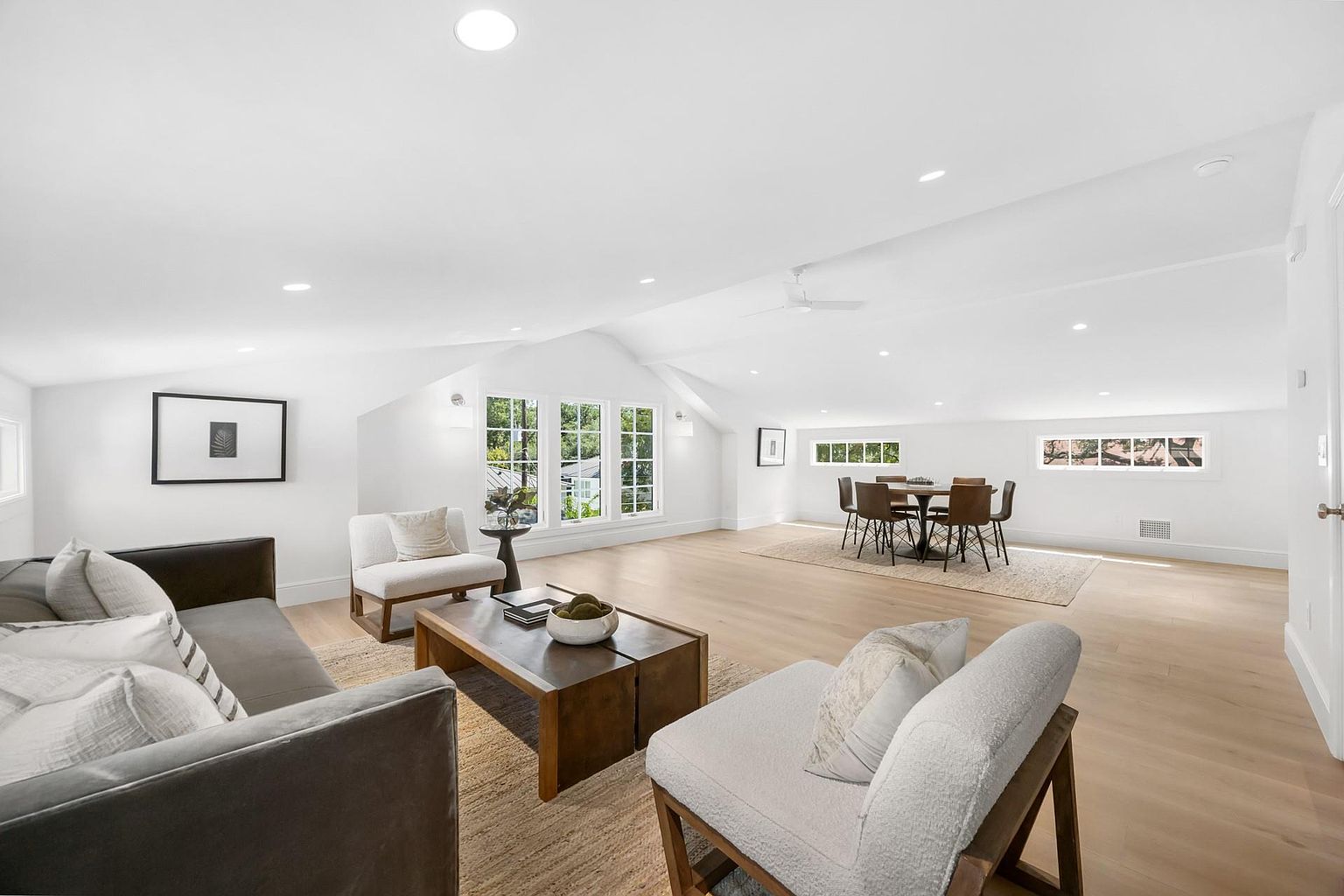 This is a bright and spacious living room with a modern aesthetic. The room features a neutral color palette, hardwood floors, and ample natural light from large windows. Furnishings include a comfortable sofa, armchairs, a coffee table, and a dining table with chairs, creating a welcoming and versatile space.