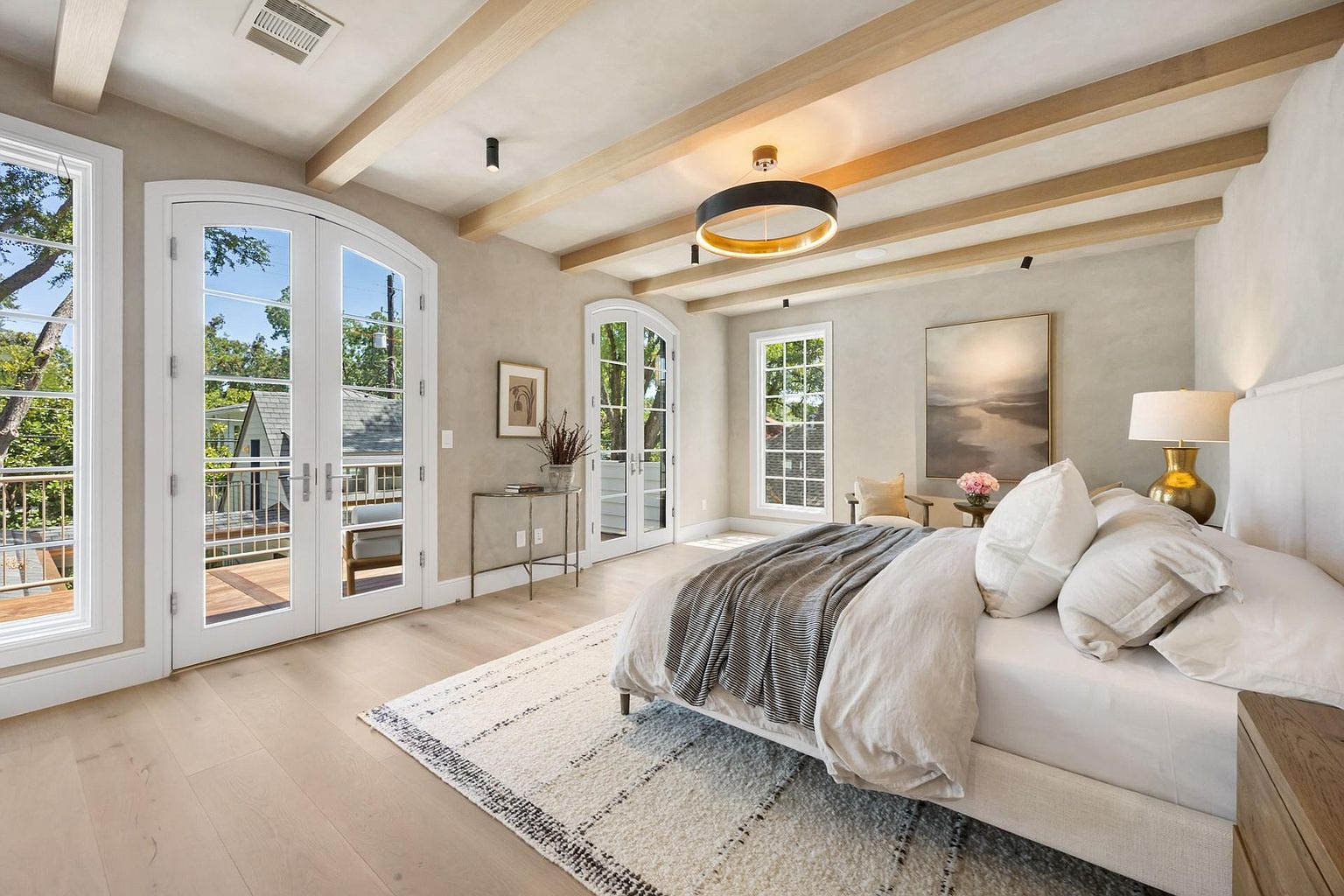 This is a bright and airy primary bedroom featuring a king-size bed with neutral linens, a textured rug, and exposed wooden beams on the ceiling. Large windows and French doors provide ample natural light and access to a balcony. The room is decorated in a sophisticated, minimalist style with a focus on natural materials and a calming color palette.