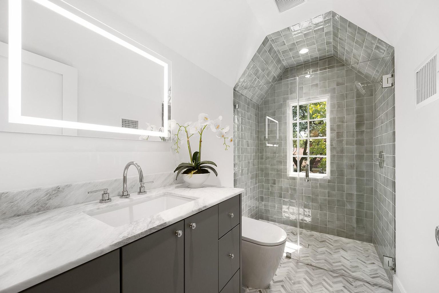 This is a well-lit primary bathroom featuring a gray vanity with a marble countertop, a modern sink, and a large, illuminated mirror. The shower area is enclosed with glass and tiled in gray, with a window providing natural light. The floor is tiled in a herringbone pattern, adding a touch of elegance.