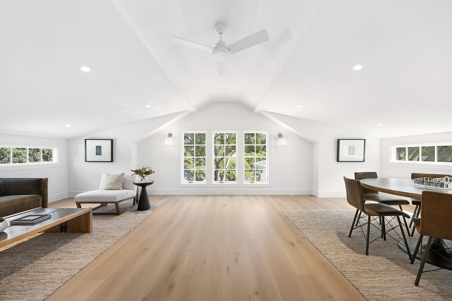 This is a bright and airy living room featuring a vaulted ceiling with a ceiling fan and recessed lighting. Large windows provide ample natural light and views of the outdoors. The room is furnished with a sofa, chaise lounge, coffee table, dining table and chairs, and is decorated with neutral colors and simple artwork, creating a clean and modern aesthetic.