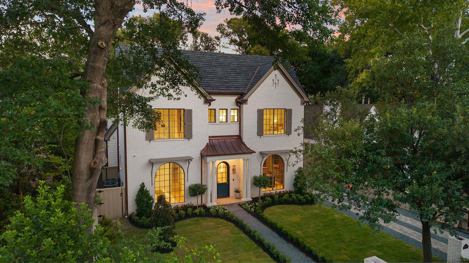 This is a front exterior view of a two-story white brick house with a dark gray roof. The house features arched windows, dark shutters, and a covered entryway with copper accents. The front yard is well-manicured with a stone pathway leading to the front door, and mature trees frame the house, creating a sense of established elegance and curb appeal.