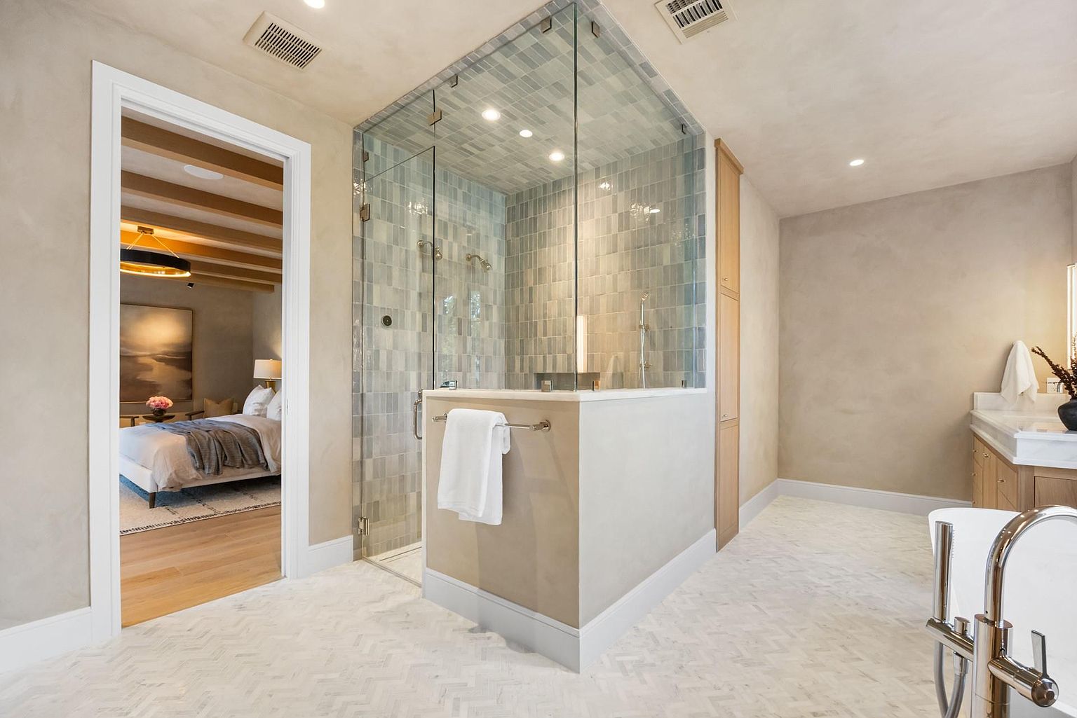 This is a bright and airy primary bathroom featuring a glass-enclosed shower with light blue tiling and a built-in bench. The flooring is a herringbone pattern, and the walls are a neutral tone. An open doorway leads to the primary bedroom, creating a sense of flow and spaciousness.