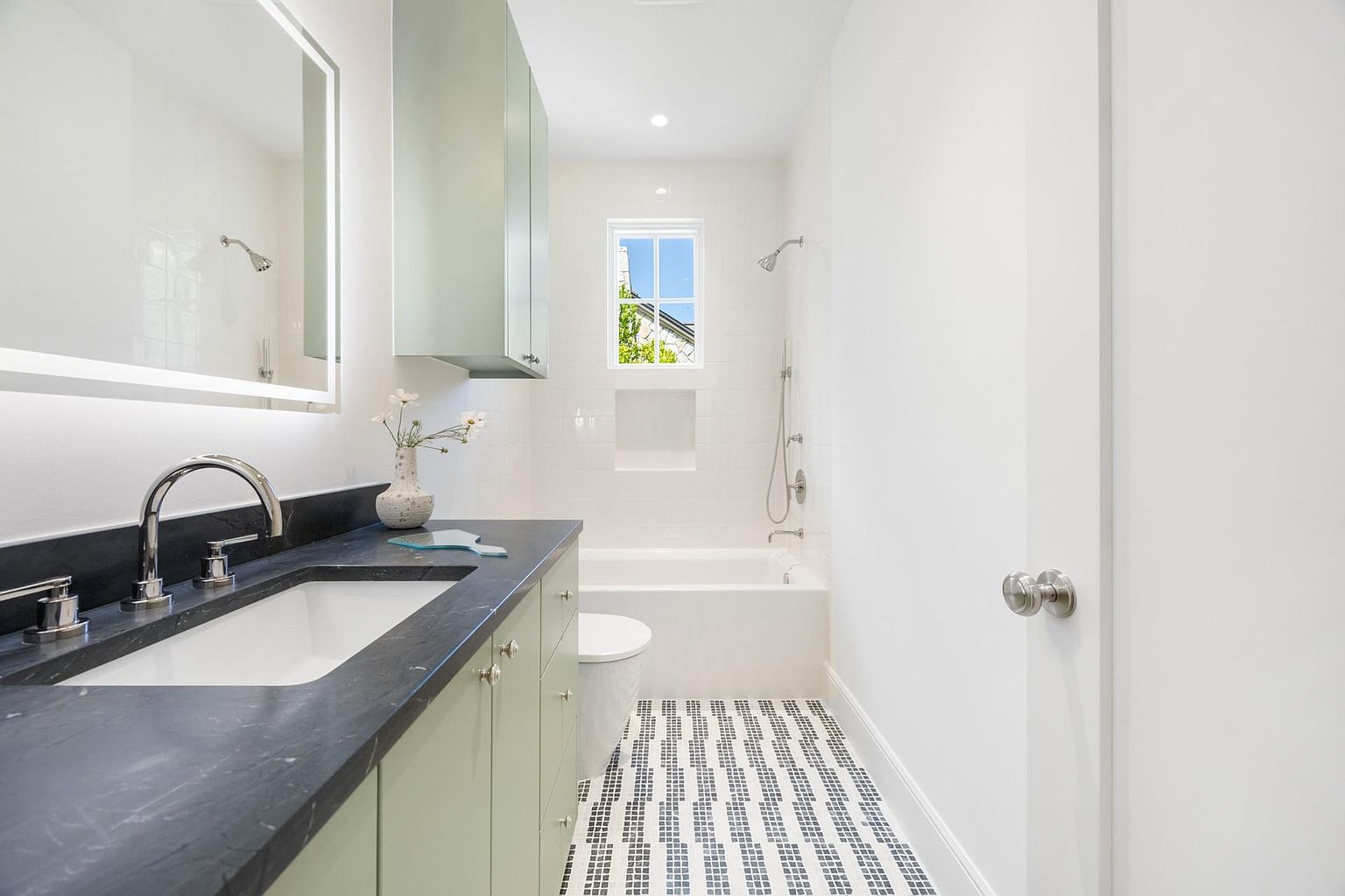 This is a well-lit bathroom featuring a vanity with a dark countertop and light green cabinetry. A large, illuminated mirror hangs above the sink, and a combination tub and shower is visible in the background. The floor is tiled with a geometric pattern, and the overall aesthetic is clean and modern.