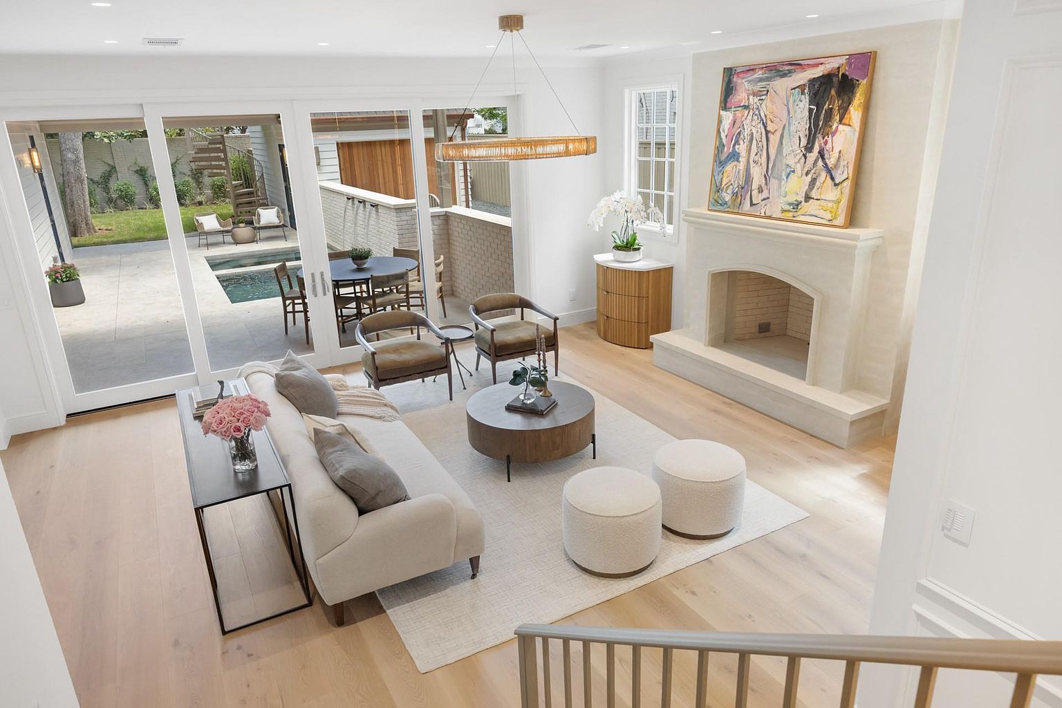 This is an interior shot of a bright and airy living room. The room features a neutral color palette with light wood floors, a white sofa, and a stone fireplace. Large sliding glass doors lead to an outdoor patio and pool area, creating a seamless indoor-outdoor living experience.