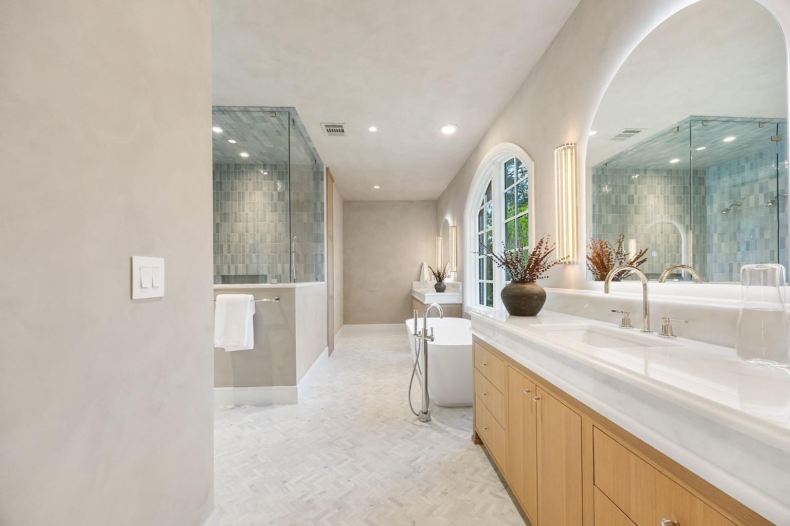 This is a bright and luxurious primary bathroom featuring a double vanity with a white countertop and light wood cabinetry. An arched mirror with integrated lighting adds a modern touch, while a freestanding bathtub and glass-enclosed shower offer spa-like amenities. The herringbone tile flooring and neutral color palette create a serene and elegant atmosphere.