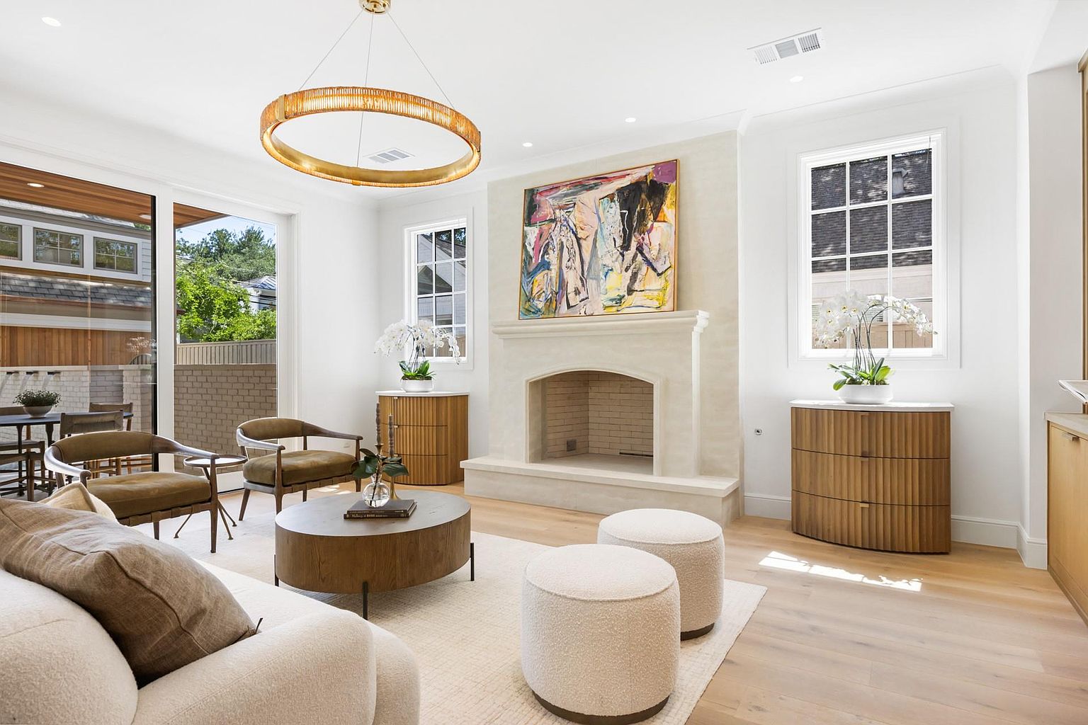 This is an interior shot of a living room featuring a neutral color palette with white walls and light wood flooring. The room is furnished with a cream-colored sofa, two olive-toned armchairs, a round wooden coffee table, and two white ottomans. A large abstract painting hangs above a fireplace, and a modern gold chandelier adds a touch of elegance. Large windows provide natural light, enhancing the bright and airy feel of the space.