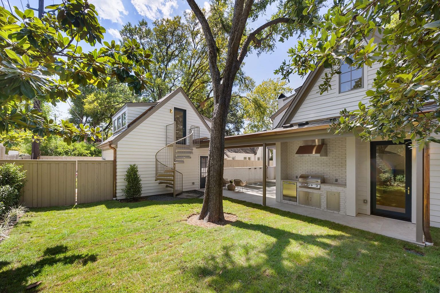 This is a rear exterior view of a home showcasing a well-maintained yard and garden area. The house features white siding, a spiral staircase leading to an upper level, and an outdoor kitchen area with a built-in grill. A mature tree provides shade, and the overall impression is one of a private and inviting outdoor living space.