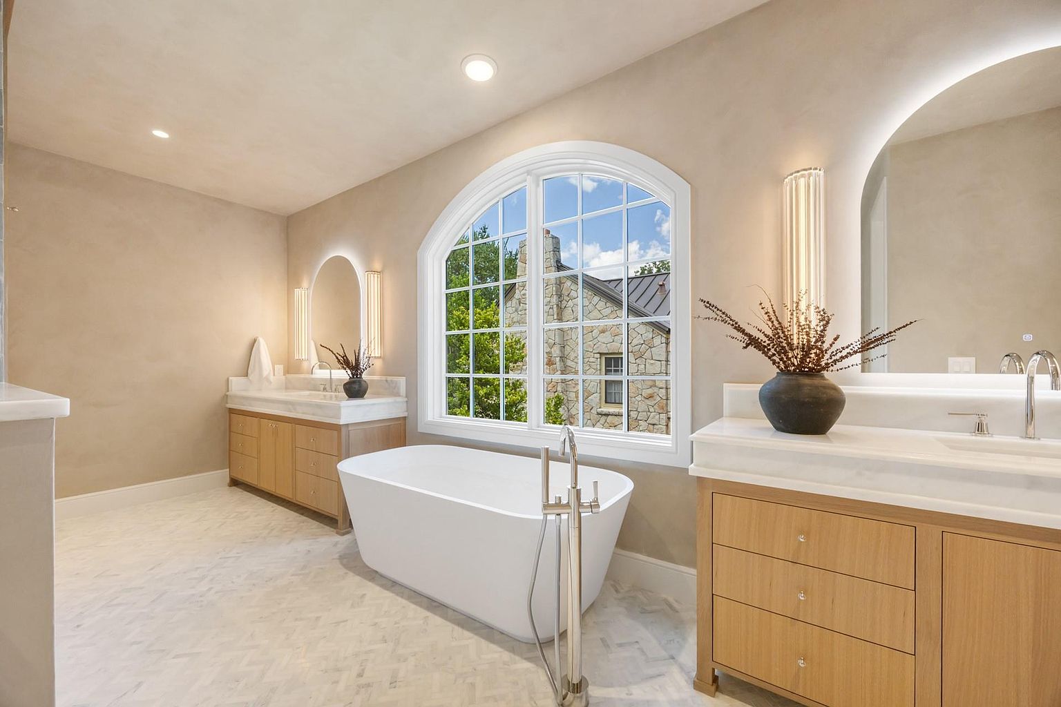 This is a luxurious primary bathroom featuring a freestanding white bathtub positioned beneath a large arched window that offers a view of a stone house and greenery. The room includes two separate vanity areas with light wood cabinetry, marble countertops, and illuminated mirrors. The flooring is a light-colored herringbone pattern, contributing to the bathroom's elegant and serene atmosphere.