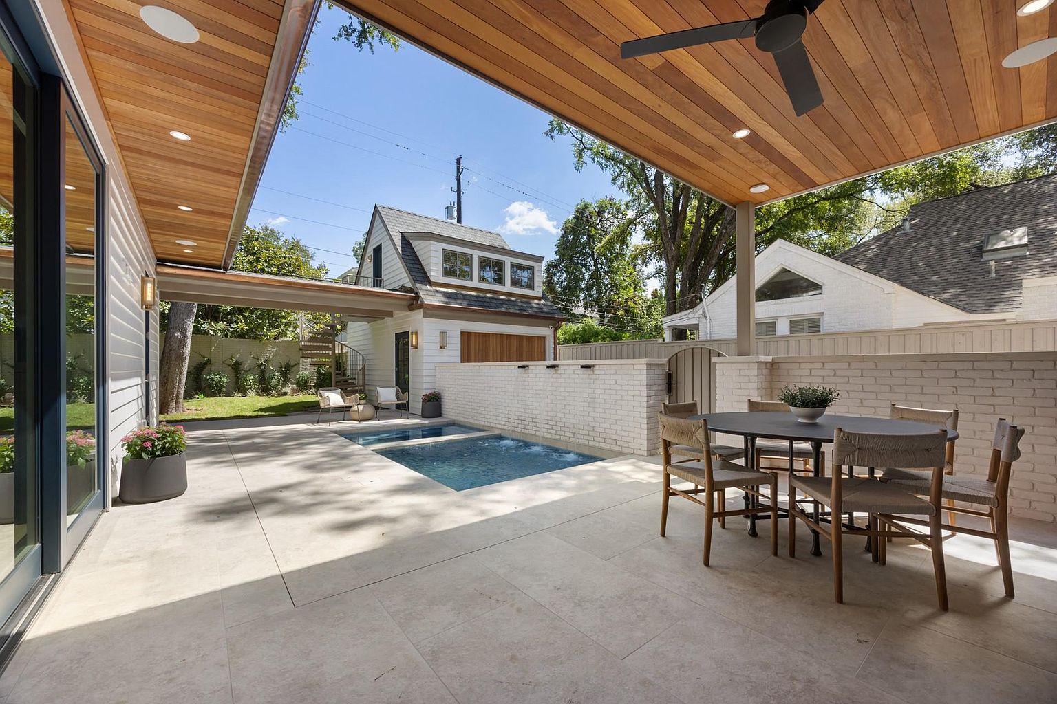 This image showcases a beautifully designed outdoor patio area, featuring a modern pool or spa, a dining set with a round table and woven chairs, and a stylish wooden ceiling with recessed lighting. The patio is paved with large, light-colored tiles, and a white brick wall adds privacy and architectural interest. The overall impression is one of luxury and relaxation, perfect for outdoor entertaining.